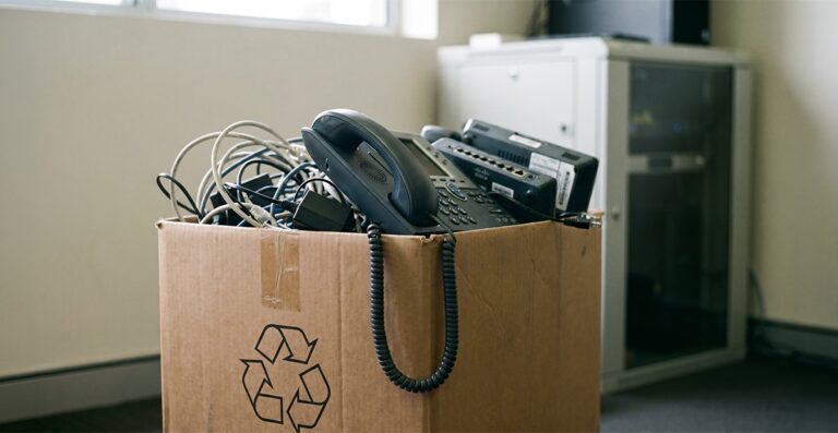 A Cisco IP desk phone handset hanging out of a cardboard recycling box of retired office phone equipment in an Australian small business, illustrating business phone e-waste.